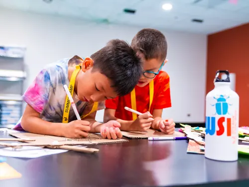 Two campers working on drawings confer at a lab table