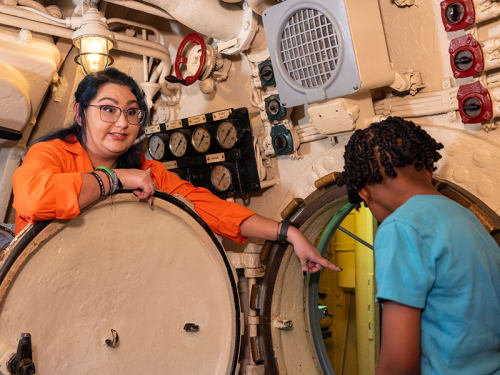A Museum guide points to an open hatch as a guest looks into it