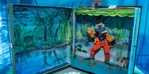 A miniature frog puppet dressed in a bright suit waves from inside a handmade stage that closes up like a book.