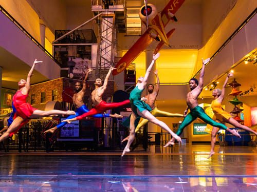 Leaping dancers mid-air in front of a hanging airplane