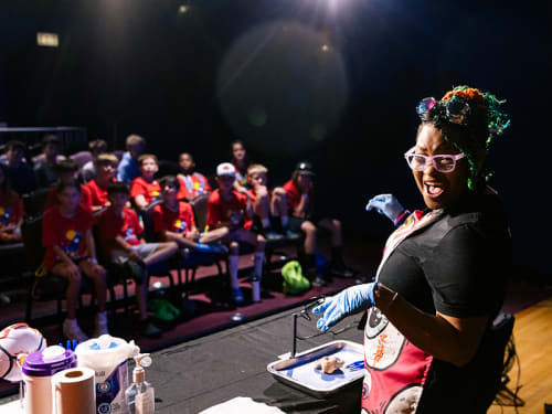 Smiling Museum facilitator turned to camera while presenting a tray with an eyeball to a group of youths