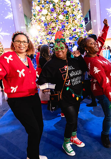 Partygoers in holiday sweaters pose in front of a large holiday tree