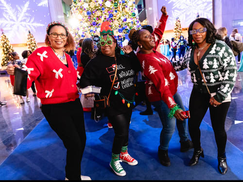 Partygoers in holiday sweaters pose in front of a large holiday tree