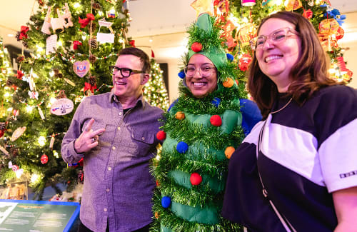 Three adults - one wearing a holiday tree costume - pose in front of actual holiday trees