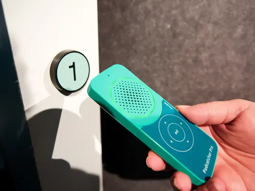 Hand holding a green audio device with speaker near a wall button marked "1"
