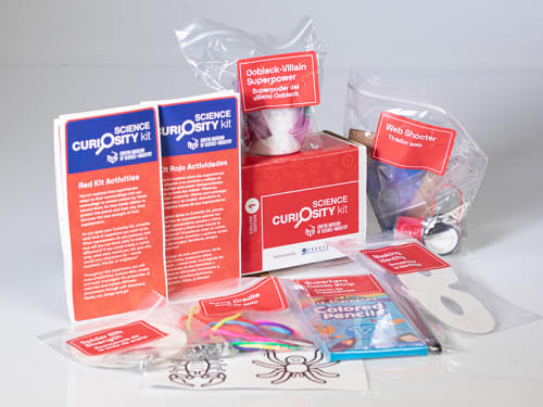 The contents of the "Red" Science Curiosity Kit are displayed featuring activities like Masking Your Identity, Web Shooter, Cat’s Cradle, Spider Silk Strength, Oobleck-Villain Superpowers, and Comic Strips