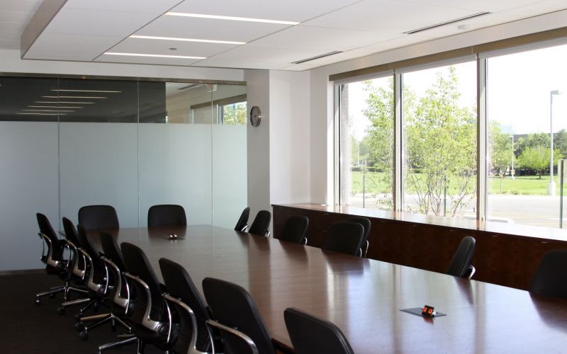 A large meeting room with a long, wood table and many chairs, alongside floor-to-wall glass windows.