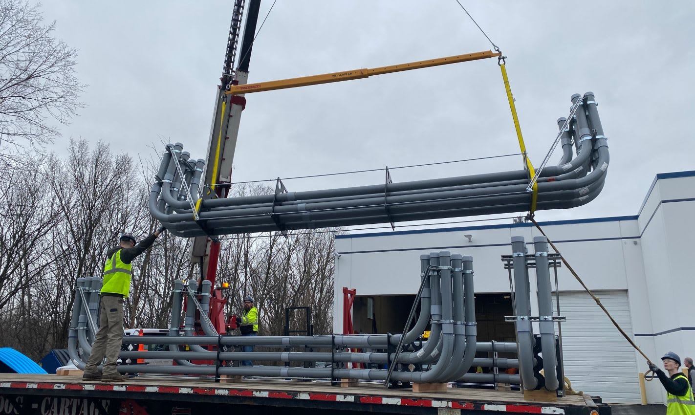 A crane lifting prefabricated electrical multi-conduit racks at a construction site.