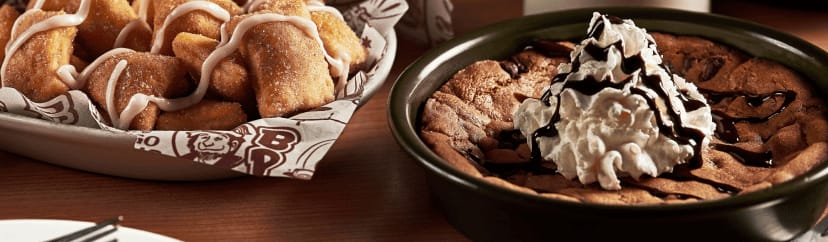 A basket of sweet dough bites drizzled in icing and a deep-dish chocolate chip cookie skillet topped with whipped cream and chocolate sauce.