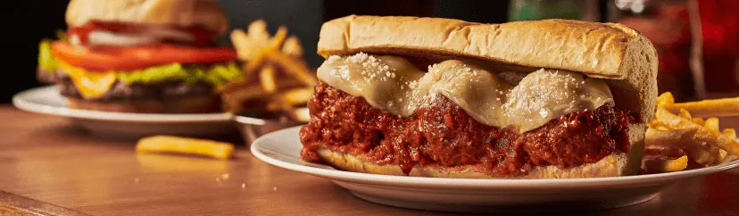 A Beggars Pizza meatball sub sandwich served with fries, with a burger and fries in the background.