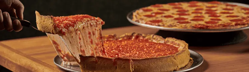 A steamy slice of deep-dish pizza from Beggars Pizza is lifted with a pie server. A pepperoni pizza sits in the background.