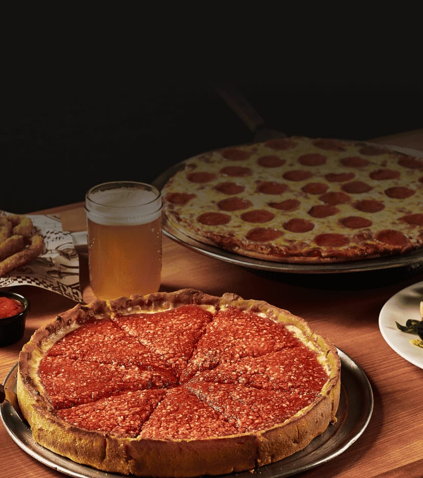 A thin-crust pepperoni pizza, a deep-dish pizza, and a salad sit on a wooden table among beer and wine glasses.