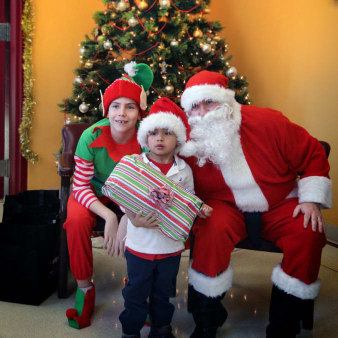 Kids being handed presents under a christmas tree next to santa and another kid in an elf suit