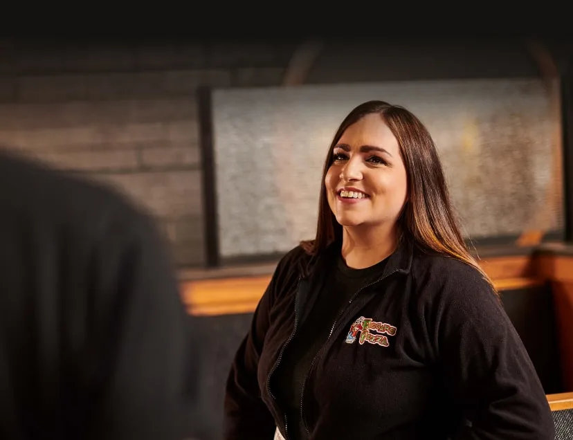 A Beggars Pizza employee wearing a branded jacket smiles at a person off camera.
