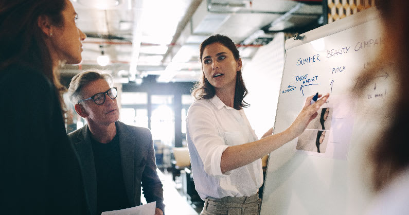 one person standing near a white board explaining a concept to two colleagues