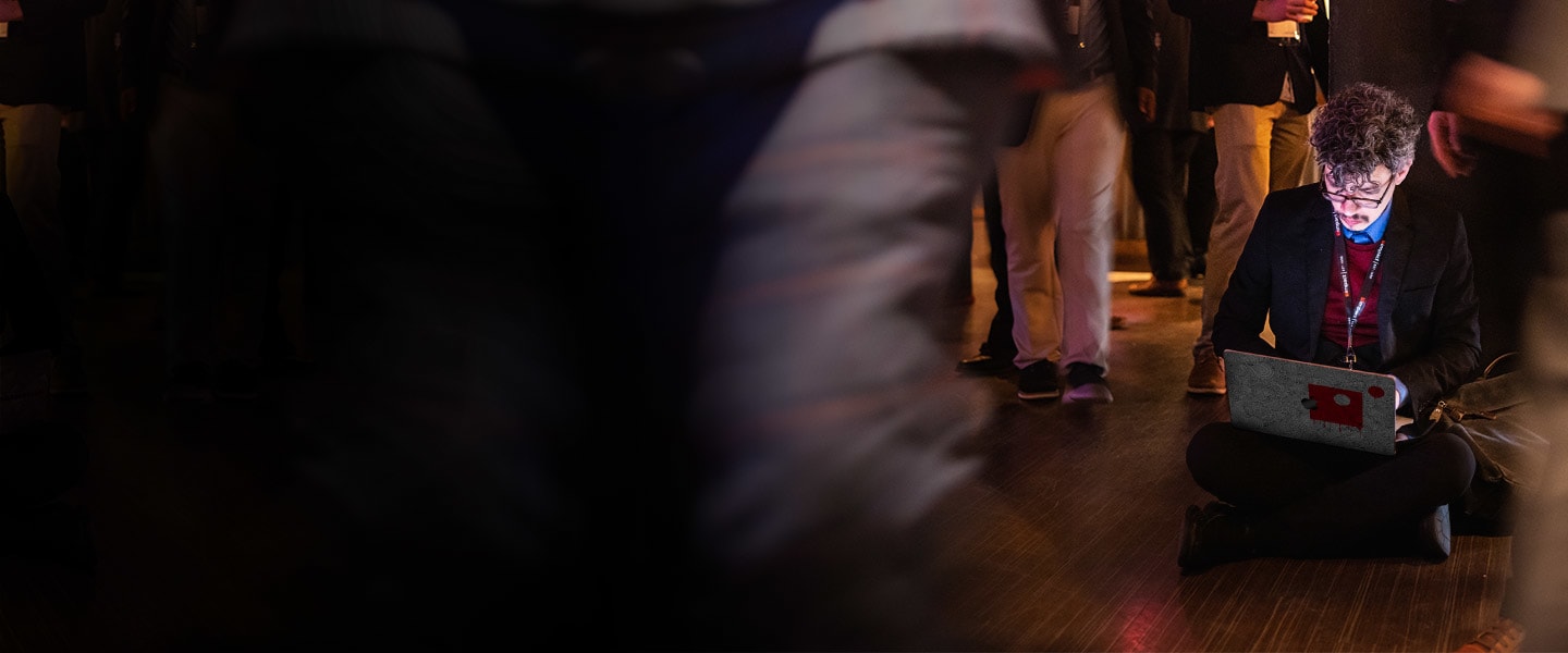 Professional sitting at a table with an Impact branded laptop, blurred figures moving about.