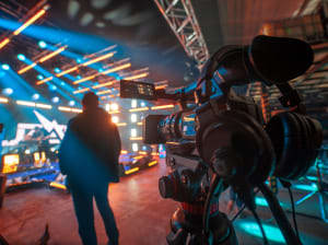 A video camera pointed at a person standing in front of a colorfully-lit studio set