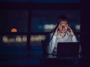 Distressed man holding his temples at a desk with his laptop open in a dark room