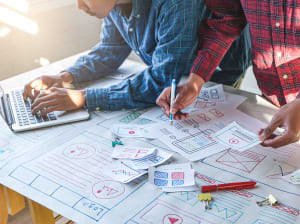 Two people at a workstation. One is working on a laptop, the other is working with a pen and hardcopy designs