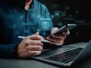 A person sitting at a laptop holding a stylus in one hand and a smartphone in the other