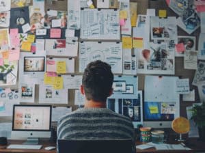 A person sitting at a work desk in front of a pin board that is full of notes and images and other brainstorming paperwork