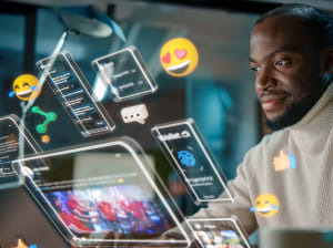 A man sits at a laptop with a variety of virtual floating displays showing various tabs he might have open working with b2b paid and social media. Also a handful of floating emojis like heart eyes and crying laughing