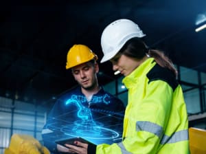 A male and female construction worker using a tablet with a digital hologram of a machine floating above the tablet.