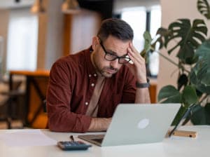A man sitting behind an open laptop holding his brow in frustration. 