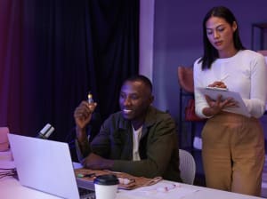 A man sits at a desk with a laptop and a marker in hand - a woman stands behind him with a notepad and pen in hand. They collaborate.