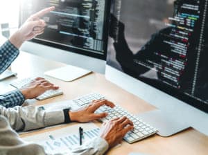 A pair of people sitting at a group of monitors writing and reviewing lines of code