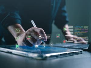 A person using a tablet pen on one screen with a laptop open next to it. There are digital icons hovering above both