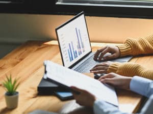 One person typing at a laptop with another showing them a report on a clipboard