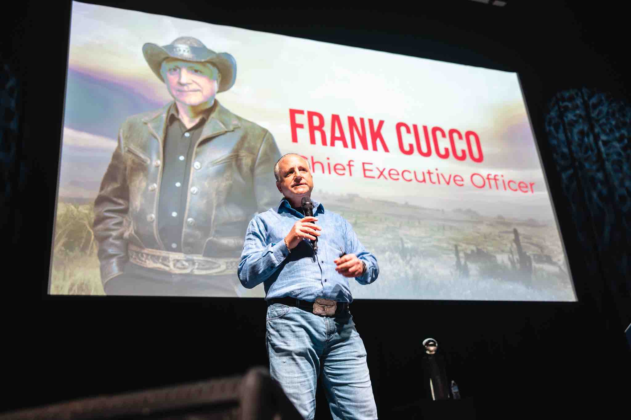 a man in jeans on stage with an an generated image of himself on a display screen behind him