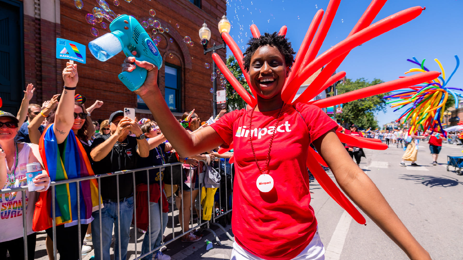 An Impact employee in a branded shirt with a dozen long balloons behind her at the Chicago Pride Parade