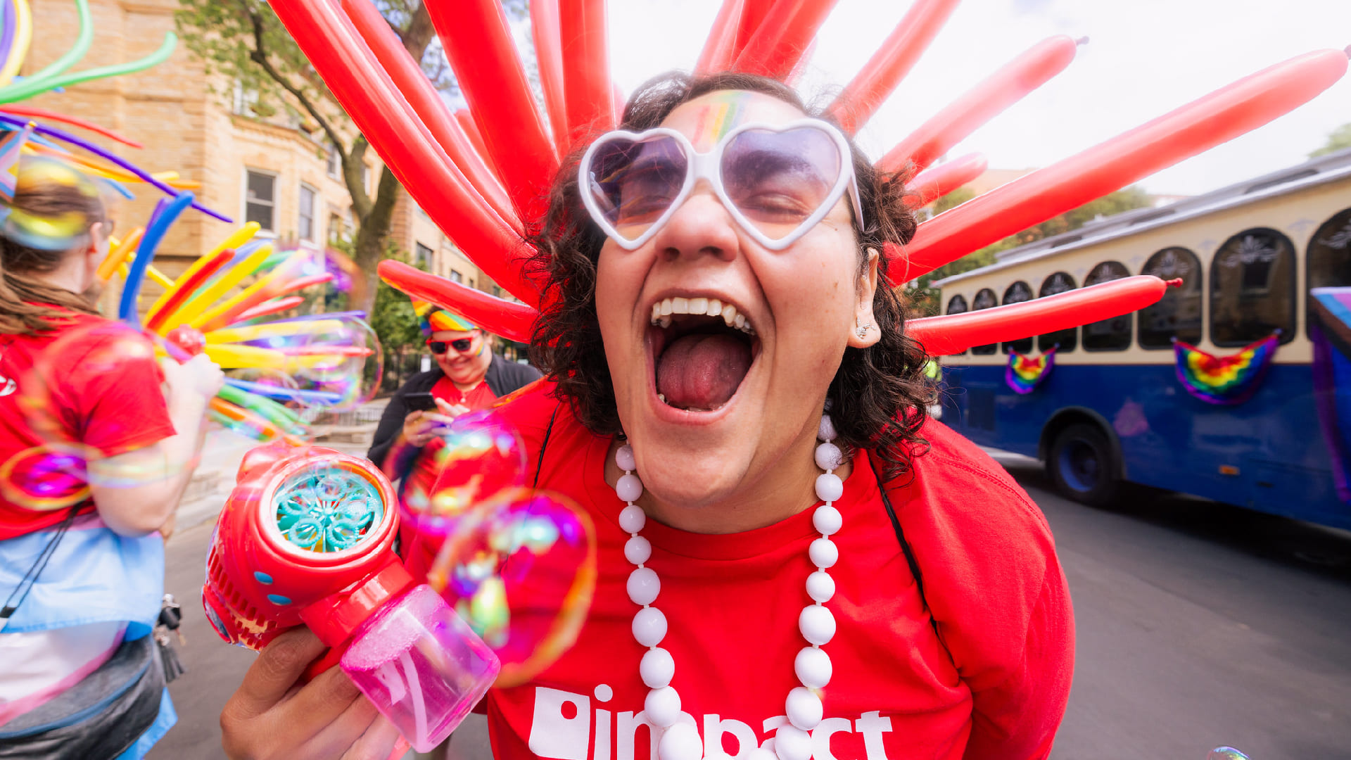 An excited woman in an Impact shirt with heart-shaped sunglasses, a bubble gun, and balloons
