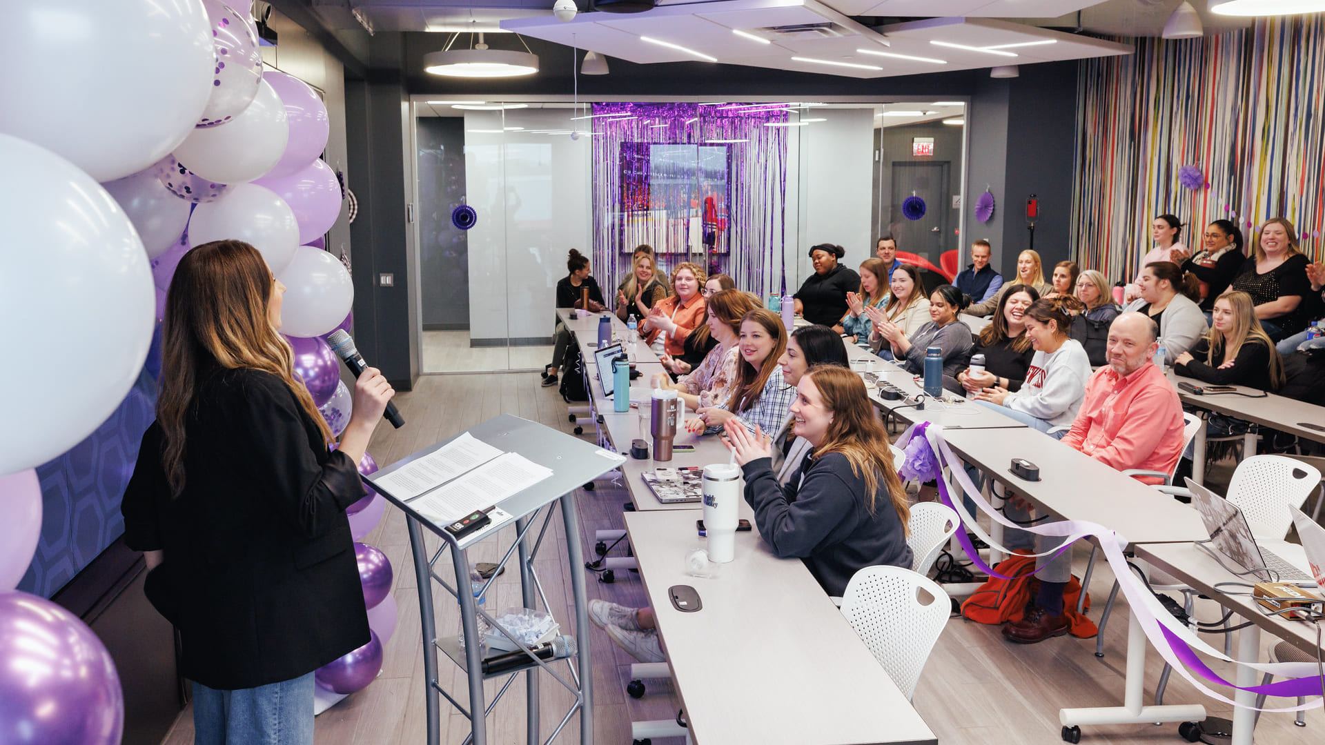 A woman with a microphone surrounded by balloons presenting to a group of people at long tables facing her